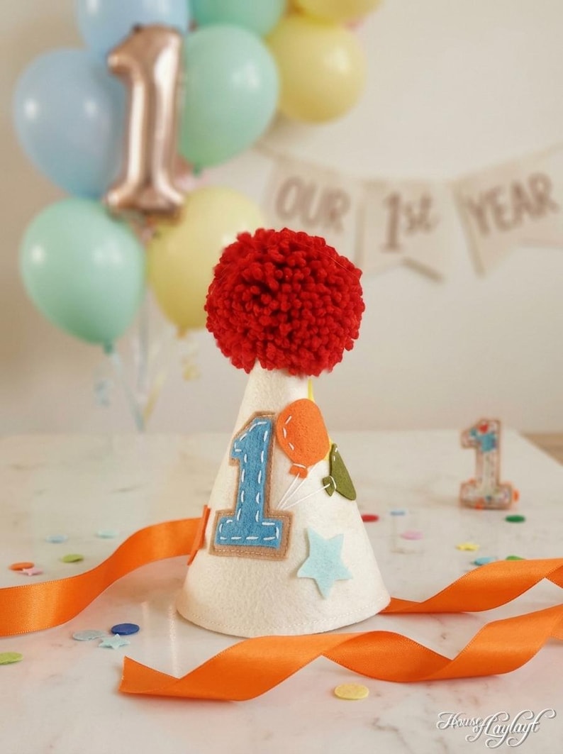 May include: A cream-colored cone-shaped party hat topped with a large red yarn pom-pom. The hat features a blue felt number one, an orange balloon, and a light blue star appliqu&eacute;. A long orange satin ribbon is attached to the base of the hat.