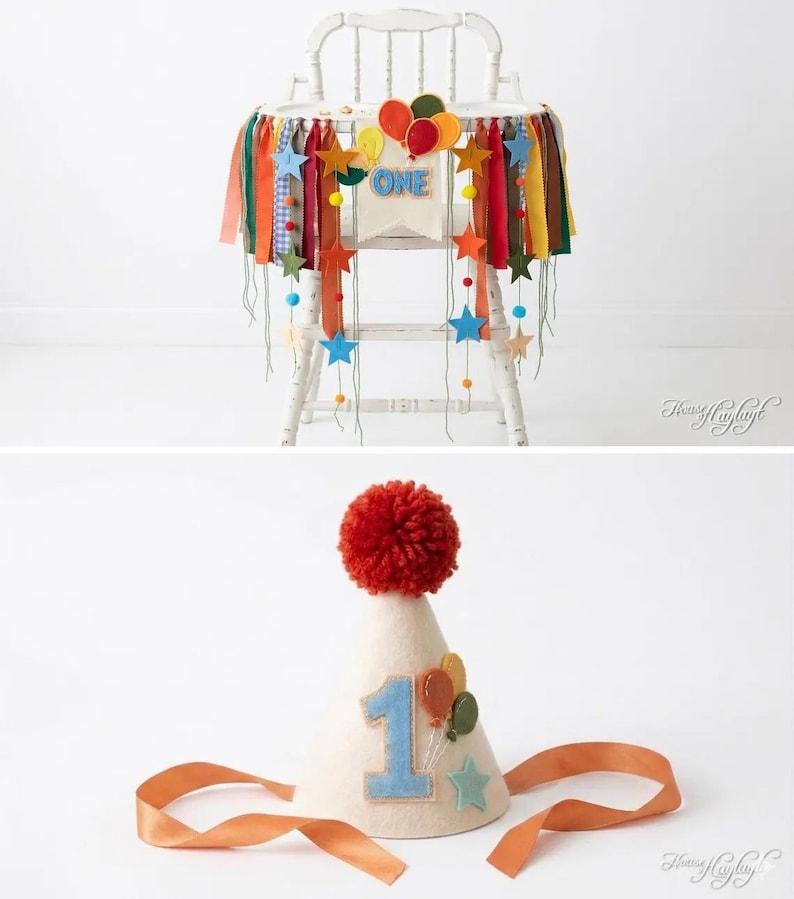 May include: A white high chair decorated with a colorful fabric banner featuring felt stars, balloons, and the word ONE, paired with a matching cone-shaped party hat topped with a red yarn pom-pom and featuring a blue number one and balloons.