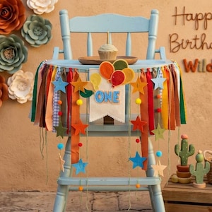 May include: A light blue high chair decorated with a fabric banner featuring the word ONE, colorful felt balloons, stars, and hanging ribbons in shades of orange, green, and blue. A small cupcake sits on a wooden plate on the chair tray.