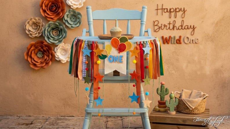 May include: A light blue high chair decorated with a fabric banner featuring the word ONE, colorful felt balloons, stars, and hanging ribbons in shades of orange, green, and blue. A small cupcake sits on a wooden plate on the chair tray.