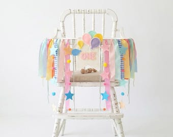 May include: A white wooden high chair decorated with a pastel-colored banner featuring fabric stars, ribbons, and felt balloons. A central sign displays the word ONE in pink letters, accented with small pom-poms and hanging star-shaped cutouts.