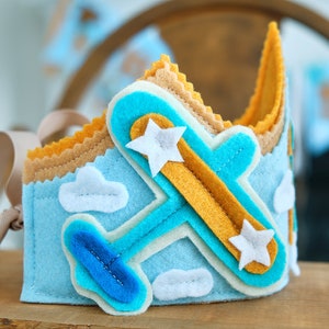 May include: A handmade felt crown in shades of blue, tan, and yellow, featuring an airplane design with white star accents and cloud details. The crown has a ribbon tie for adjustable sizing.