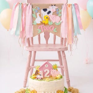 May include: A pink high chair decorated for a second birthday. The chair has a burlap banner with felt farm animals and the word "TWO". A cake with farm animal decorations sits in front of the chair, with balloons in the background.