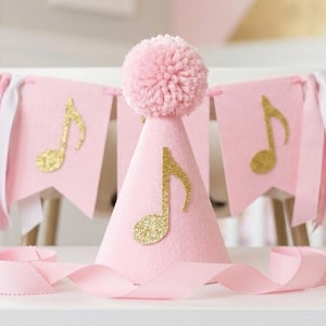 May include: A pink party hat with a gold glitter musical note and a pink pom-pom. A pink banner with gold musical notes and pink and white ribbons is behind the hat. Pink ribbon is in front.