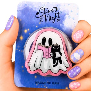 May include: A magnetic grip featuring a white ghost reading a pink book with a black cat. The grip has a pink glitter border and is on a blue background with the text "Stars at Night co."