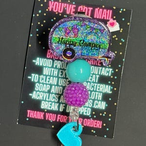 May include: A glittery purple and blue badge reel with a camper design that says "Happy Camper". The reel has a turquoise heart-shaped clip and two decorative beads. The text "YOU'VE GOT MAIL" is at the top.
