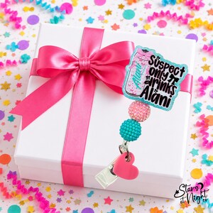 May include: A badge reel featuring a glittery acrylic topper shaped like a beverage can with the text "Suspect only drinks Alani" in black lettering. The reel includes a pink rhinestone bead, a teal textured bead, and a pink heart-shaped charm.