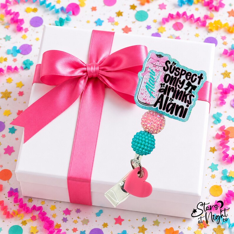 May include: A badge reel featuring a glittery acrylic topper shaped like a beverage can with the text "Suspect only drinks Alani" in black lettering. The reel includes a pink rhinestone bead, a teal textured bead, and a pink heart-shaped charm.