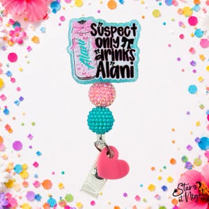 May include: A badge reel featuring a glittery light blue acrylic topper with a graphic of a pink can and the text "Suspect only drinks Alani". Below the topper are two textured beads in pink and teal, followed by a solid pink heart charm.