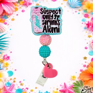 May include: A badge reel featuring a glittery light blue acrylic topper with the text Suspect only drinks Alani, a pink bow, and a can graphic. Below are two textured beads in pink and teal, followed by a solid pink heart charm and a metal clip.