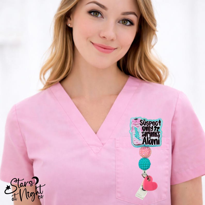 May include: A pink V-neck scrub top featuring a badge reel attached to the pocket. The badge reel has a pink beverage can graphic with the text "Suspect only drinks Alani", followed by a pink bead, a teal bead, and a pink heart charm.