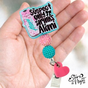 May include: A badge reel featuring a glittery teal-bordered acrylic topper with a graphic of a beverage can and the text "Suspect only drinks Alani". The reel includes a pink rhinestone bead, a teal textured bead, and a pink heart charm.