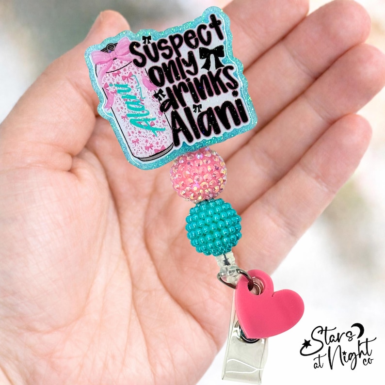 May include: A badge reel featuring a glittery teal-bordered acrylic topper with a graphic of a beverage can and the text "Suspect only drinks Alani". The reel includes a pink rhinestone bead, a teal textured bead, and a pink heart charm.