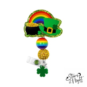 May include: A St. Patrick's Day themed badge reel featuring a green leprechaun hat, a rainbow, and a pot of gold. The badge reel has a rainbow bead, a gold rhinestone bead, and a green shamrock charm. The badge reel has a clear clip.
