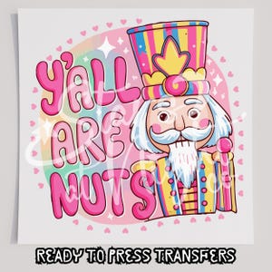 May include: A colorful graphic design featuring a nutcracker character with a striped hat and white beard, accompanied by the pink text Y'ALL ARE NUTS. The background includes a circular pastel pattern decorated with small pink hearts and star shapes.