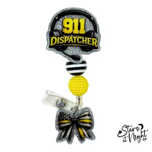 May include: A badge reel featuring a silver glitter topper with black headphones and the yellow text 911 DISPATCHER. Below are a black and white striped bead, a yellow textured bead, and a silver glitter bow charm with a thin yellow stripe.