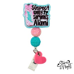 May include: A badge reel featuring a glittery light blue acrylic topper with a pink beverage can graphic and the text Suspect only drinks Alani. Below the topper are two round textured beads in pink and teal, and a solid pink heart charm.