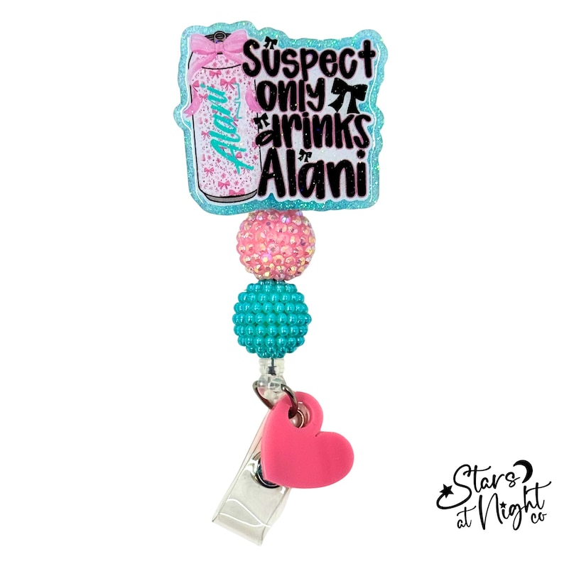 May include: A badge reel featuring a glittery light blue acrylic topper with a pink beverage can graphic and the text Suspect only drinks Alani. Below the topper are two round textured beads in pink and teal, and a solid pink heart charm.