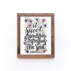 Puede incluir: Una impresión enmarcada con fondo blanco y texto negro que dice "Una dulce amistad refresca el alma." El texto está rodeado de flores coloridas y purpurina dorada. La parte inferior de la impresión dice "Proverbios 27:9".