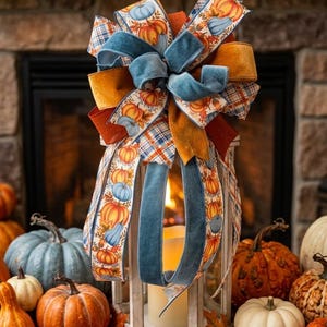 May include: A white lantern topped with a large decorative bow featuring blue velvet, orange fabric, and a pumpkin-patterned ribbon. The lantern contains a pillar candle and is surrounded by various pumpkins and scattered autumn leaves.