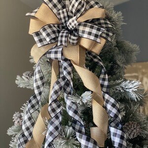 May include: A decorative Christmas tree bow featuring a combination of black and white checkered ribbon and natural burlap ribbon. The bow is placed on a flocked Christmas tree, creating a festive holiday decoration.