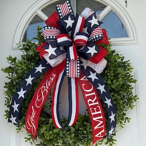 May include: A patriotic wreath with a large bow featuring red, white, and blue ribbons. The bow has star and stripe patterns, with the words "God Bless America" on the ribbons. The wreath is made of green foliage and is suitable for the Fourth of July.