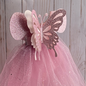 May include: A pink tulle tutu with a pink glitter butterfly on top. The butterfly has white wings and a pink body.