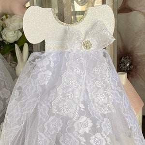 May include: White lace baby dress with a white glitter bodice and a white satin ribbon. The dress has a white bow with a rhinestone center.
