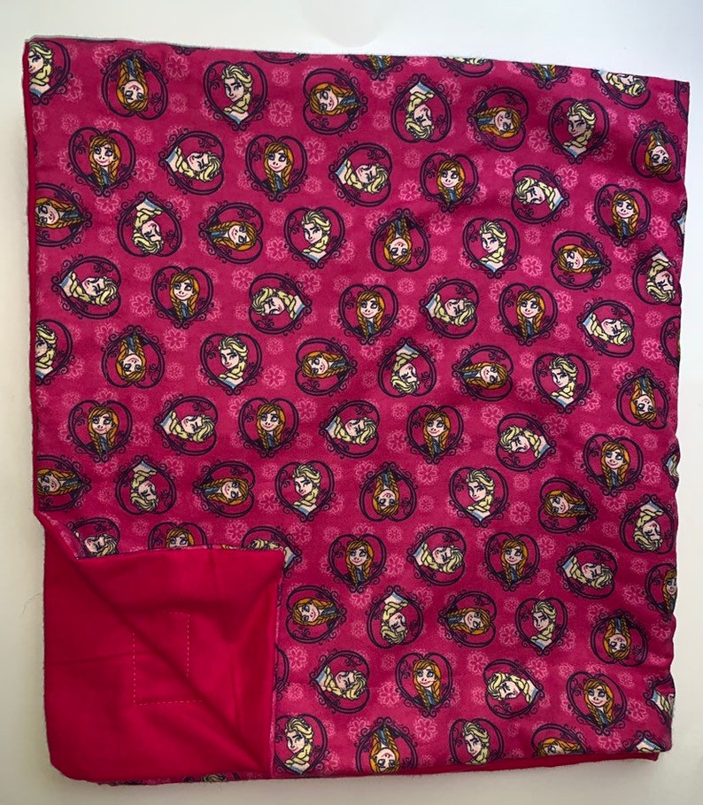 Puede incluir: Una manta de forro polar rosa con un patr&oacute;n repetido de dos chicas con cabello rubio en marcos en forma de coraz&oacute;n. Las chicas llevan vestidos azules y tienen mejillas rosadas. El fondo es un patr&oacute;n floral rosa y blanco.