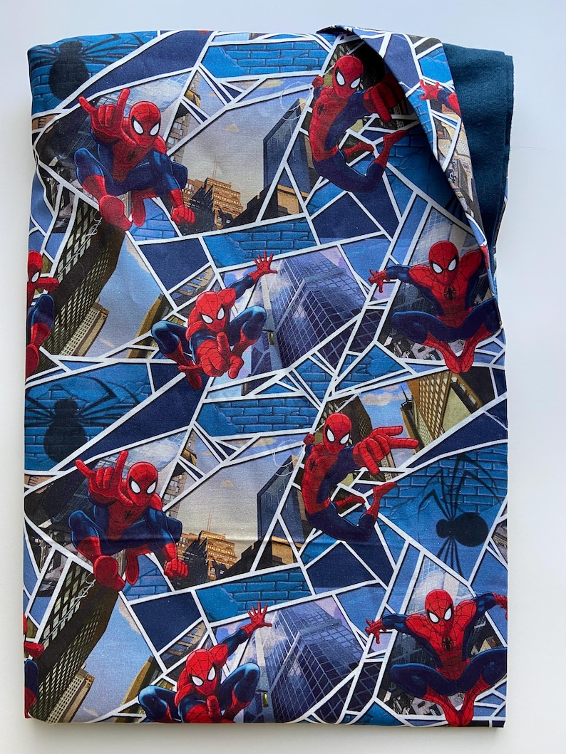 Puede incluir: Un art&iacute;culo de tela azul con un patr&oacute;n repetido de Spider-Man en poses de acci&oacute;n. El dise&ntilde;o incorpora paisajes urbanos y formas geom&eacute;tricas en tonos de azul, rojo y blanco. Tambi&eacute;n es visible una silueta de ara&ntilde;a negra.