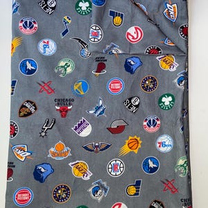 May include: Gray fabric with a pattern of NBA team logos, including the Lakers, Celtics, and Bulls. The logos are in various colors and sizes, creating a sporty design. The fabric is folded.