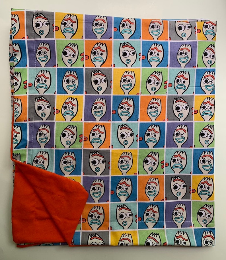 Pode incluir: Uma manta de tecido multicolorida com um padr&atilde;o repetido de um rosto de personagem de desenho animado. O personagem tem um rosto branco com olhos grandes, uma boca vermelha e uma forma de garfo para a cabe&ccedil;a. A manta tem um forro laranja s&oacute;lido.