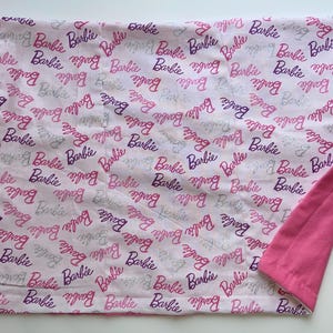 May include: A pink and white blanket featuring the word "Barbie" in a repeating pattern. The word is written in a cursive font in shades of pink, purple, and gray. The reverse side of the blanket is solid pink.