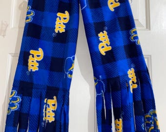 May include: A blue and navy plaid scarf with the word "Pitt" in yellow. The scarf has fringe at the ends and is hanging on a white hanger against a white door. The scarf is made of a soft material.