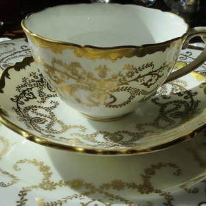 May include: A white teacup and saucer set with gold floral detailing and gold trim. The set is placed on a matching serving tray, also with gold floral patterns. The teacup has a scalloped rim.