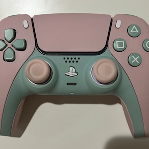 May include: A two-tone wireless game controller in pink and green. The controller features a pink faceplate and buttons, with green accents on the directional pad, triggers, and grips. The PlayStation logo is centered on the controller.