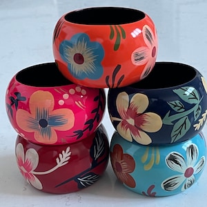 May include: Set of five colorful wooden napkin rings with floral designs. The rings are stacked in a pyramid shape, with the top ring being orange, the next ring pink, the next navy blue, the next red, and the bottom ring blue.