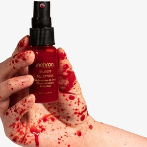 May include: A hand covered in red fake blood holds a bottle of Mehron Blood Splatter. The bottle is labelled with the product name and instructions in English, French, Spanish, and German.