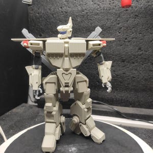 Può includere: Una action figure di robot articolato beige e grigio con dettagli rossi e blu. Il robot ha un design dettagliato con più giunture e pannelli, in piedi su una superficie scura.