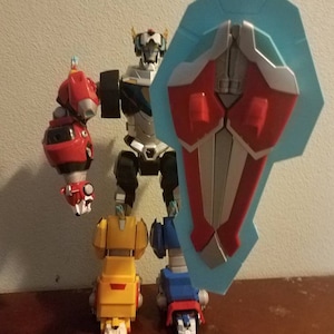 May include: A Voltron action figure, a robot made up of five lion mecha. The figure is mainly silver, red, yellow, and blue, with a light blue shield. The robot is standing on a brown surface.