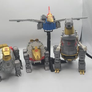 Pode incluir: Quatro figuras de ação vintage dos Transformers em modo robô e dinossauro. As figuras são prateadas, douradas e vermelhas com alguns detalhes pretos. As figuras estão colocadas sobre uma superfície branca.