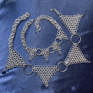 May include: A set of silver chainmail jewelry, including a necklace, two bracelets, and two triangular pieces. The pieces are made of small, interlocking metal rings and feature spikes and large circular rings. The jewelry is displayed on a blue surface.