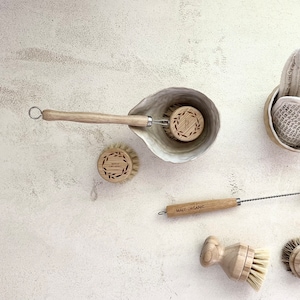 May include: A set of wooden dish brushes and a white cotton dishcloth in a ceramic bowl. The brushes have natural bristles and wooden handles. The dishcloth is folded and placed in a ceramic bowl. The items are arranged on a white textured surface.