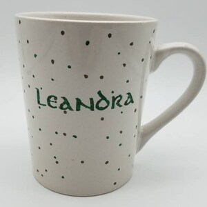 May include: White ceramic mug with a green and brown polka dot pattern. The mug has the name "Leandra" written in green on the front.