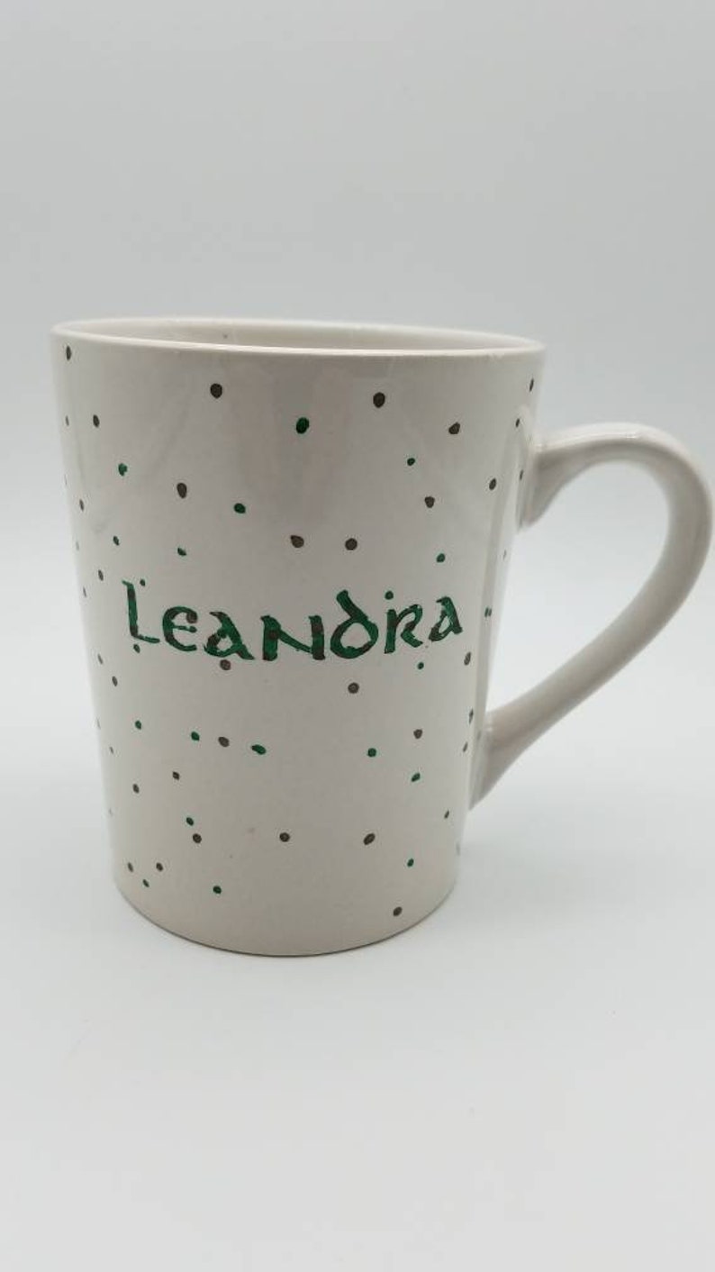May include: White ceramic mug with a green and brown polka dot pattern. The mug has the name "Leandra" written in green on the front.