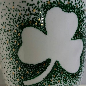 May include: White ceramic mug with a green shamrock design. The shamrock is outlined in white and the background is covered in green and gold dots.