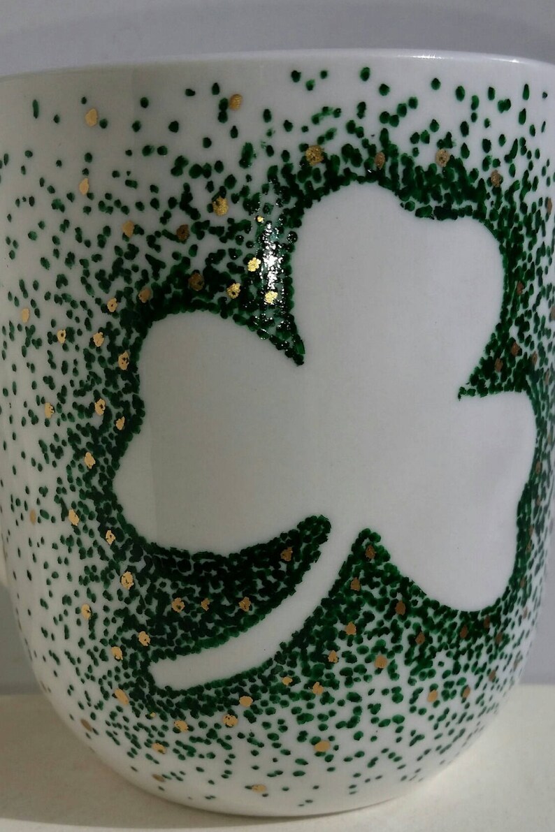 May include: White ceramic mug with a green shamrock design. The shamrock is outlined in white and the background is covered in green and gold dots.
