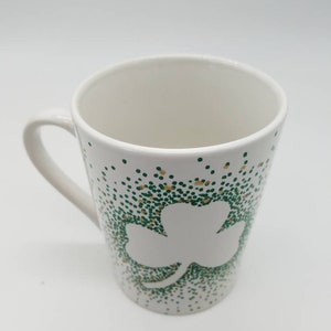 May include: White ceramic mug with a green and gold shamrock design. The shamrock is outlined in white and the background is covered in green and gold dots.