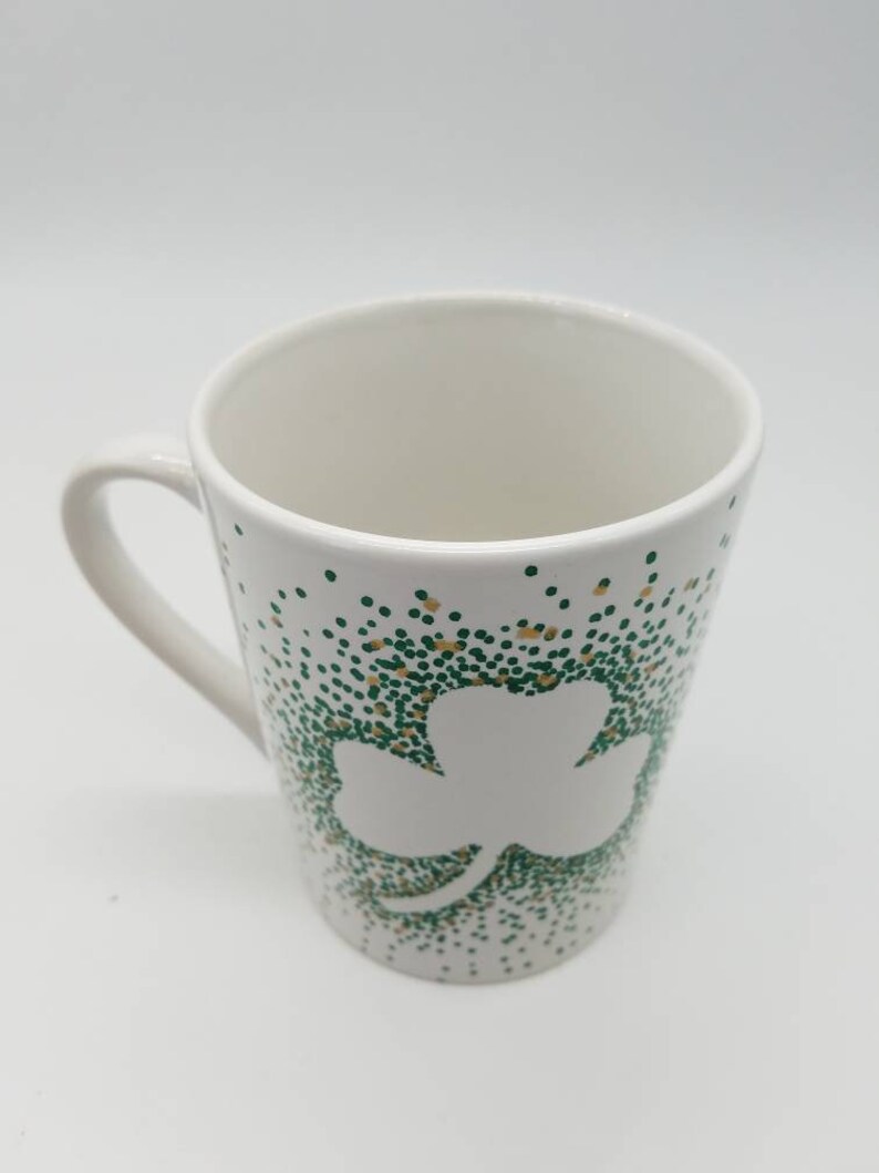 May include: White ceramic mug with a green and gold shamrock design. The shamrock is outlined in white and the background is covered in green and gold dots.