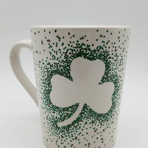 May include: White ceramic mug with a green shamrock design. The shamrock is made of green dots and the background is covered in green and brown dots.